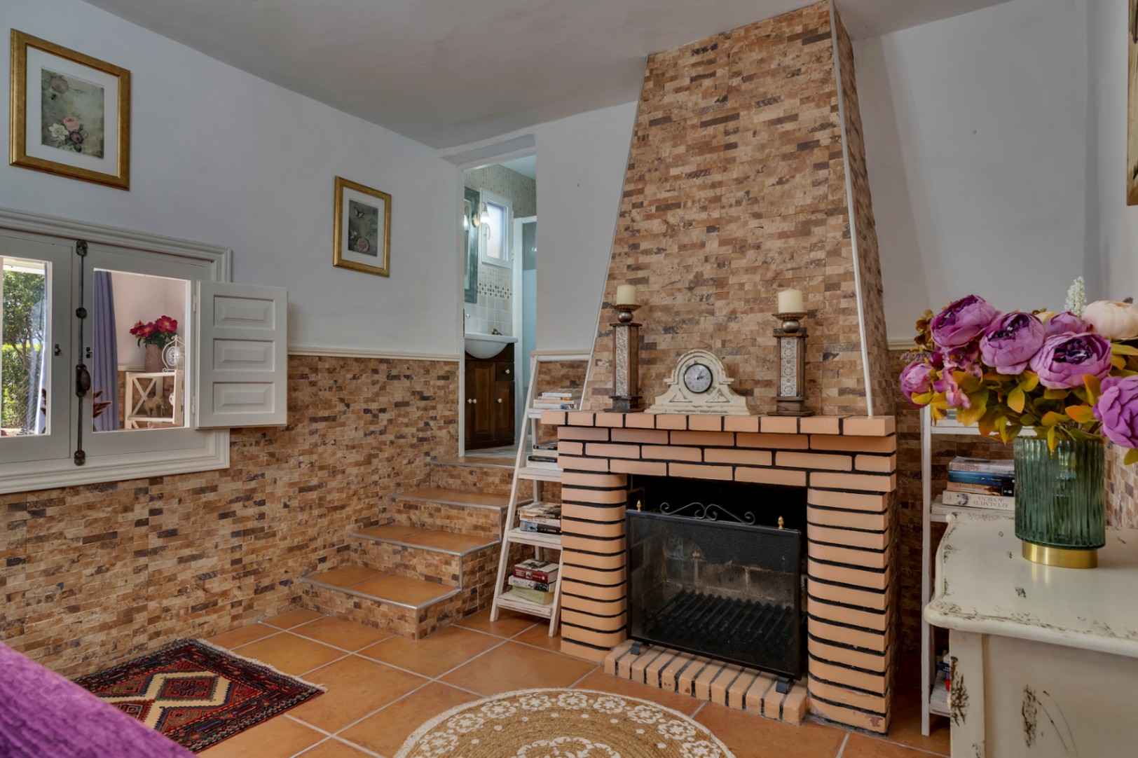Salón con chimenea de obra, paredes revestidas en piedra y suelos de gres. Una pequeña escalera conecta con el baño, aportando un diseño rústico y funcional.