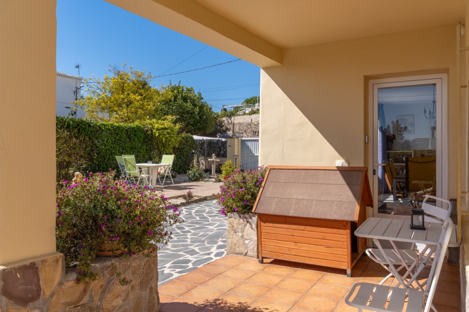 Terraza cubierta con suelos cerámicos y acceso al jardín, que cuenta con senderos de piedra natural, vegetación mediterránea y zona de comedor exterior.