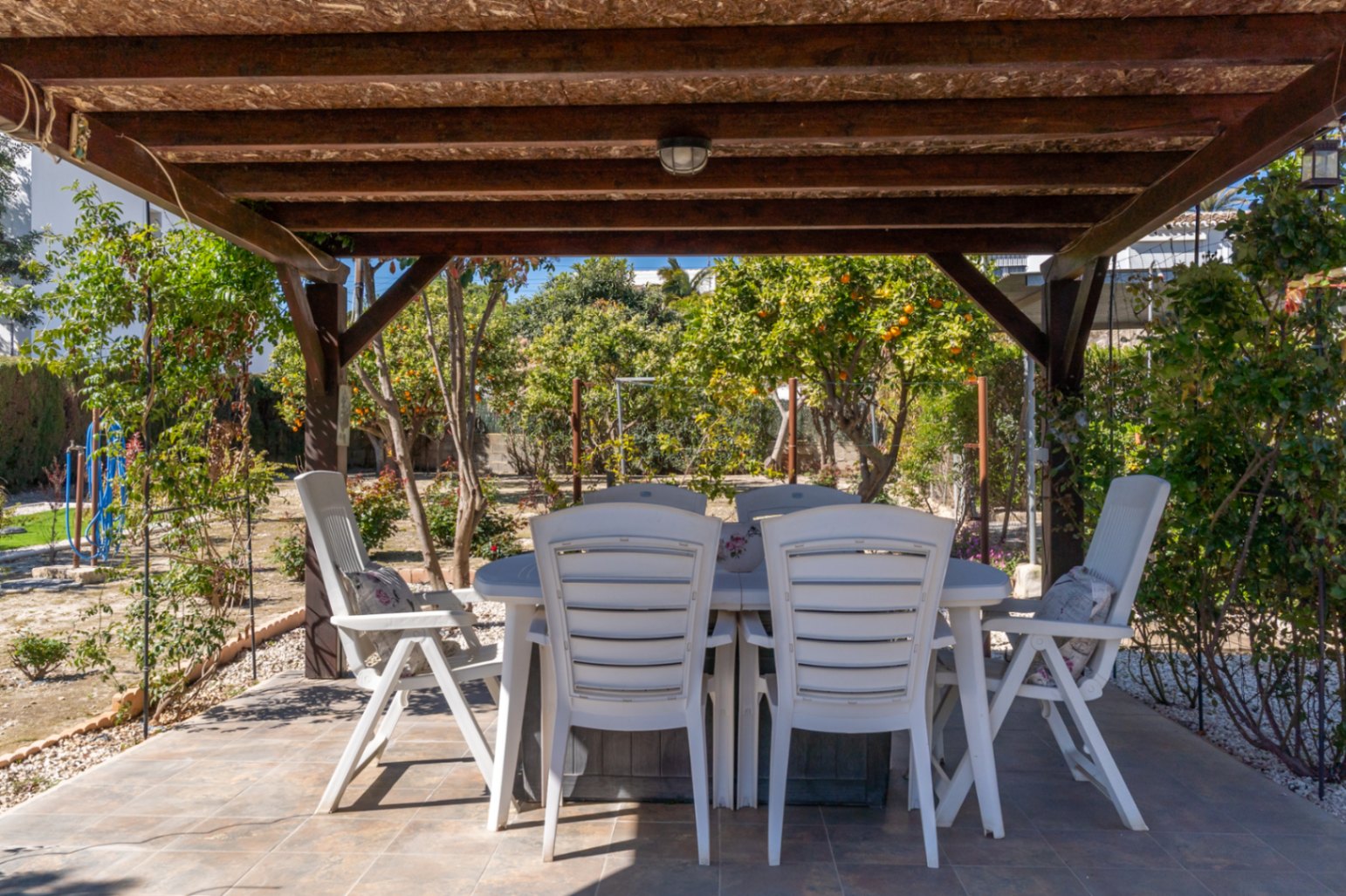 Terraza exterior cubierta por una pérgola de madera, equipada con mobiliario de comedor y vistas directas al jardín con árboles frutales.