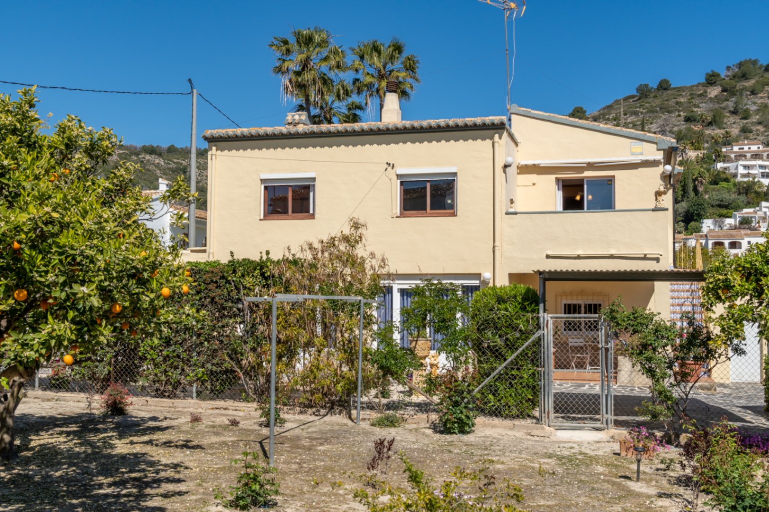 Vista exterior de la fachada sur de la villa, destacando el jardín privado con árboles frutales y la distribución en dos plantas independientes bajo el cielo despejado.