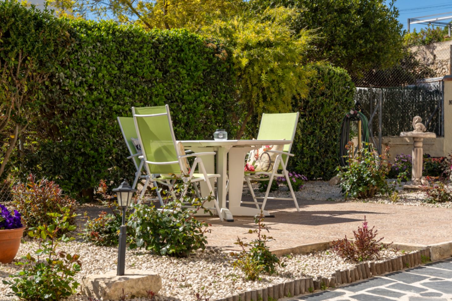 Zona de estar exterior con mesa blanca y sillas verdes sobre pavimento de piedra, rodeada de setos altos y un jardín mediterráneo con grava y plantas ornamentales.