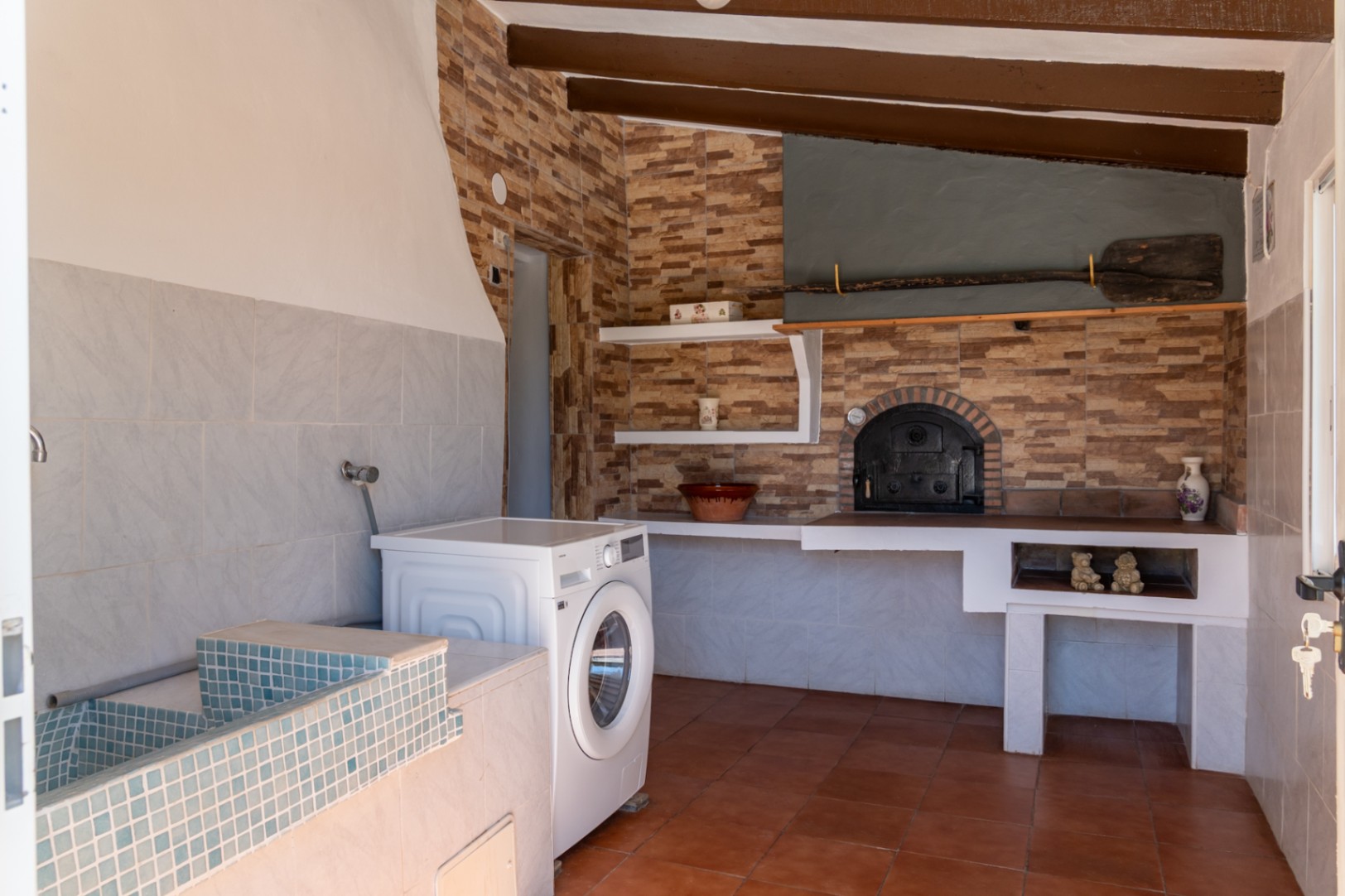 Zona de lavandería y cocina rústica equipada con lavadero de mosaico azul, lavadora y un horno de leña tradicional bajo vigas de madera vista y suelos de terracota.