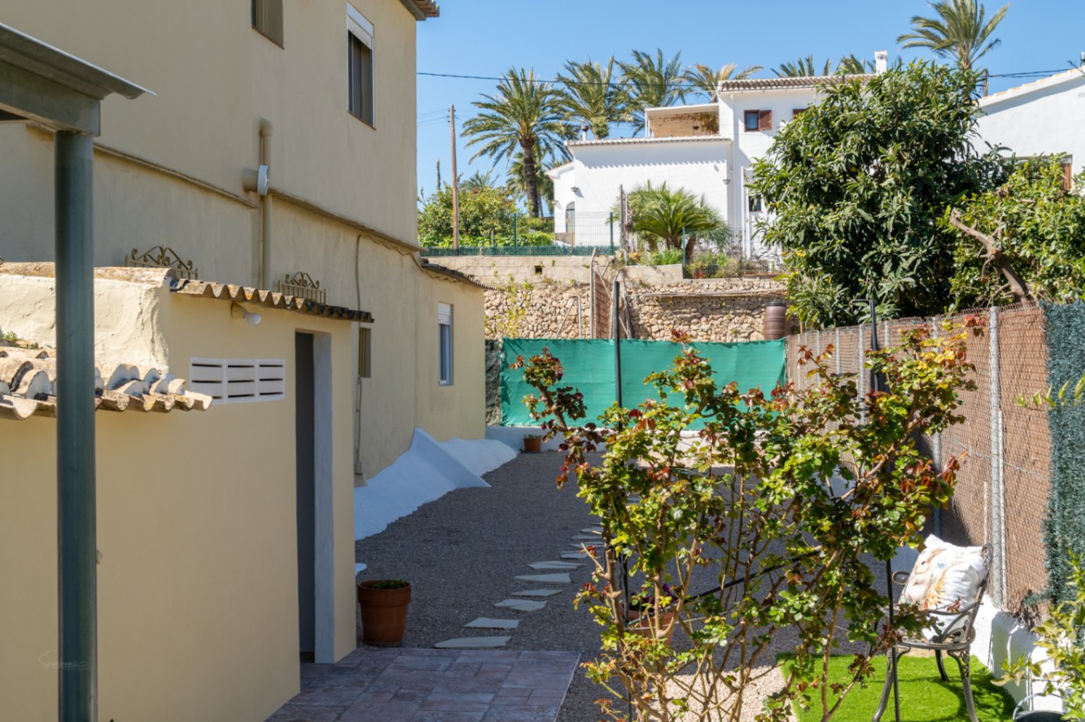 Zona exterior lateral con camino de grava y piedras de paso, vegetación mediterránea y acceso a un anexo funcional bajo el sol de Jávea.