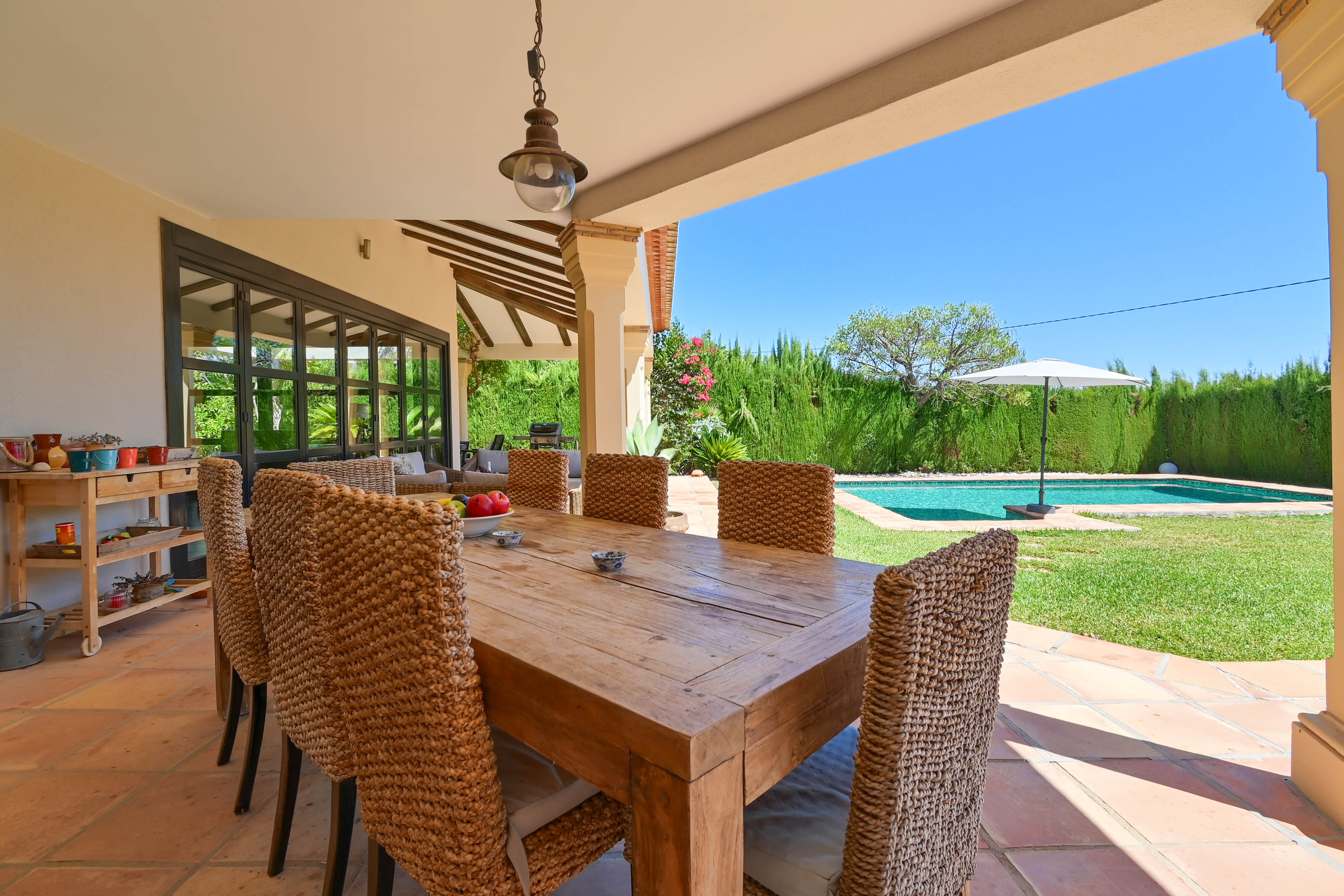 Comedor exterior cubierto con mesa de madera maciza y sillas de ratán con vistas a la piscina y al jardín.