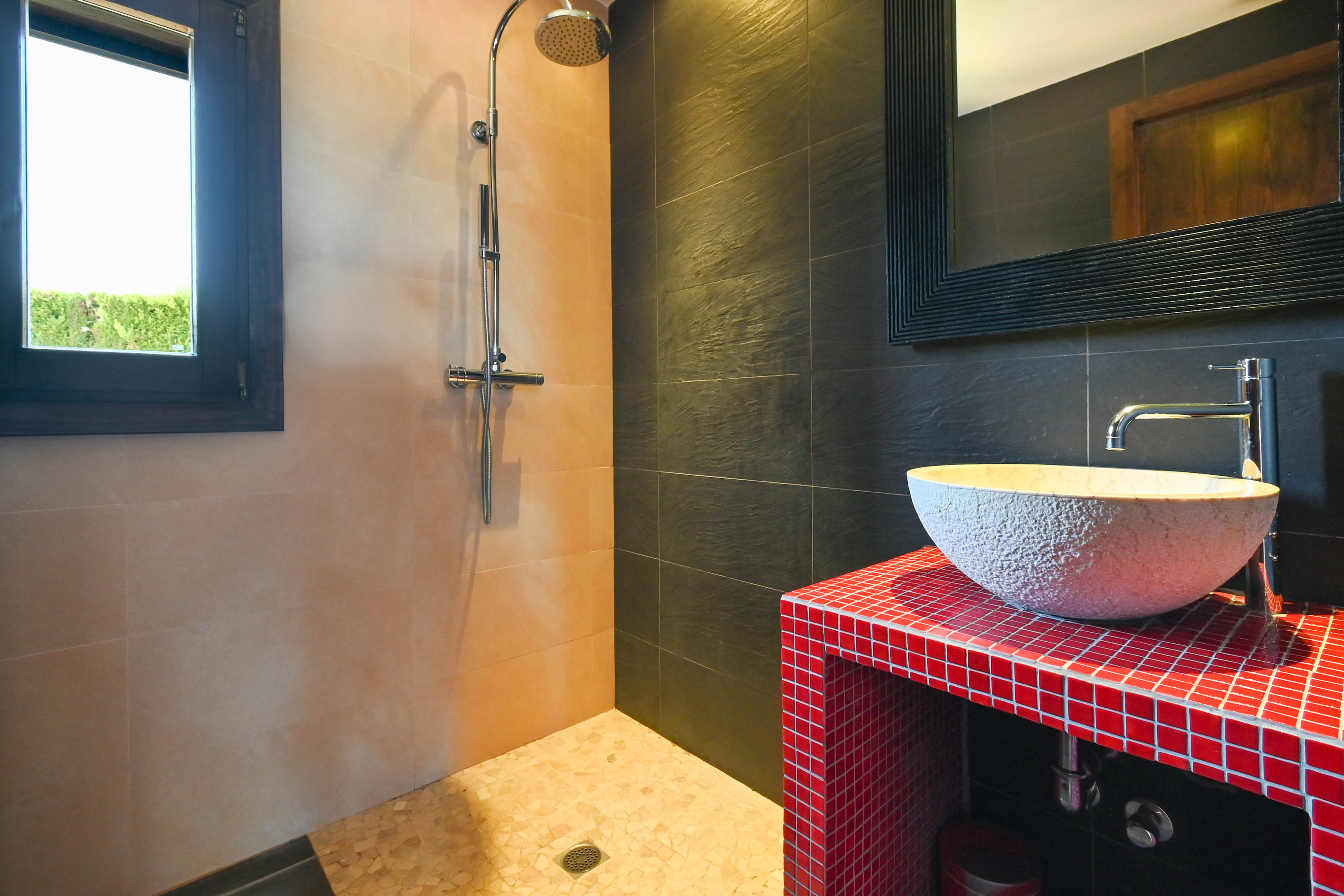 Detalle de baño con ducha, lavabo rústico sobre encimera roja de mosaico y azulejos en tonos neutros y oscuros.