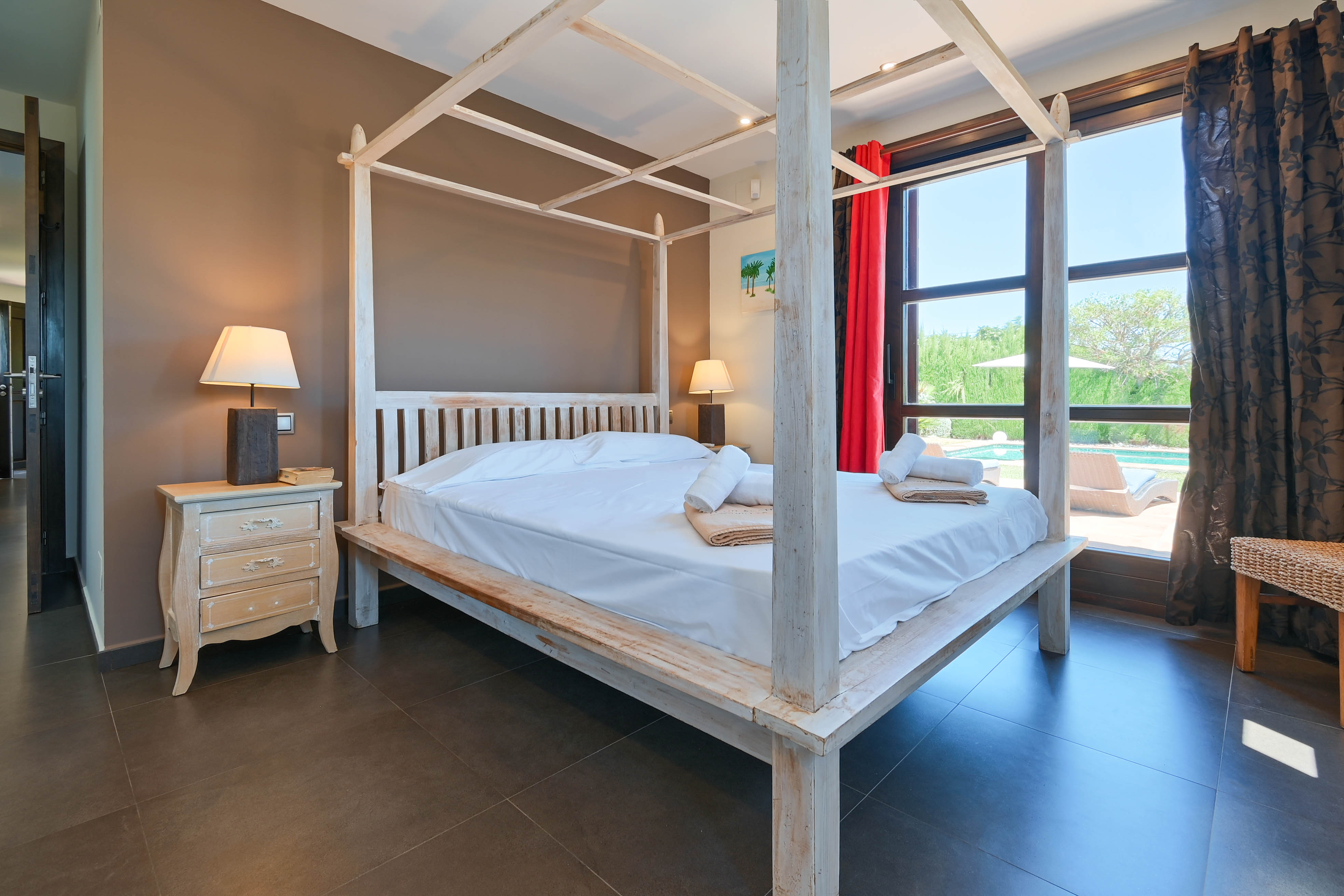 Dormitorio con cama con dosel de madera blanca, mesita de noche rústica y vistas a la piscina.
