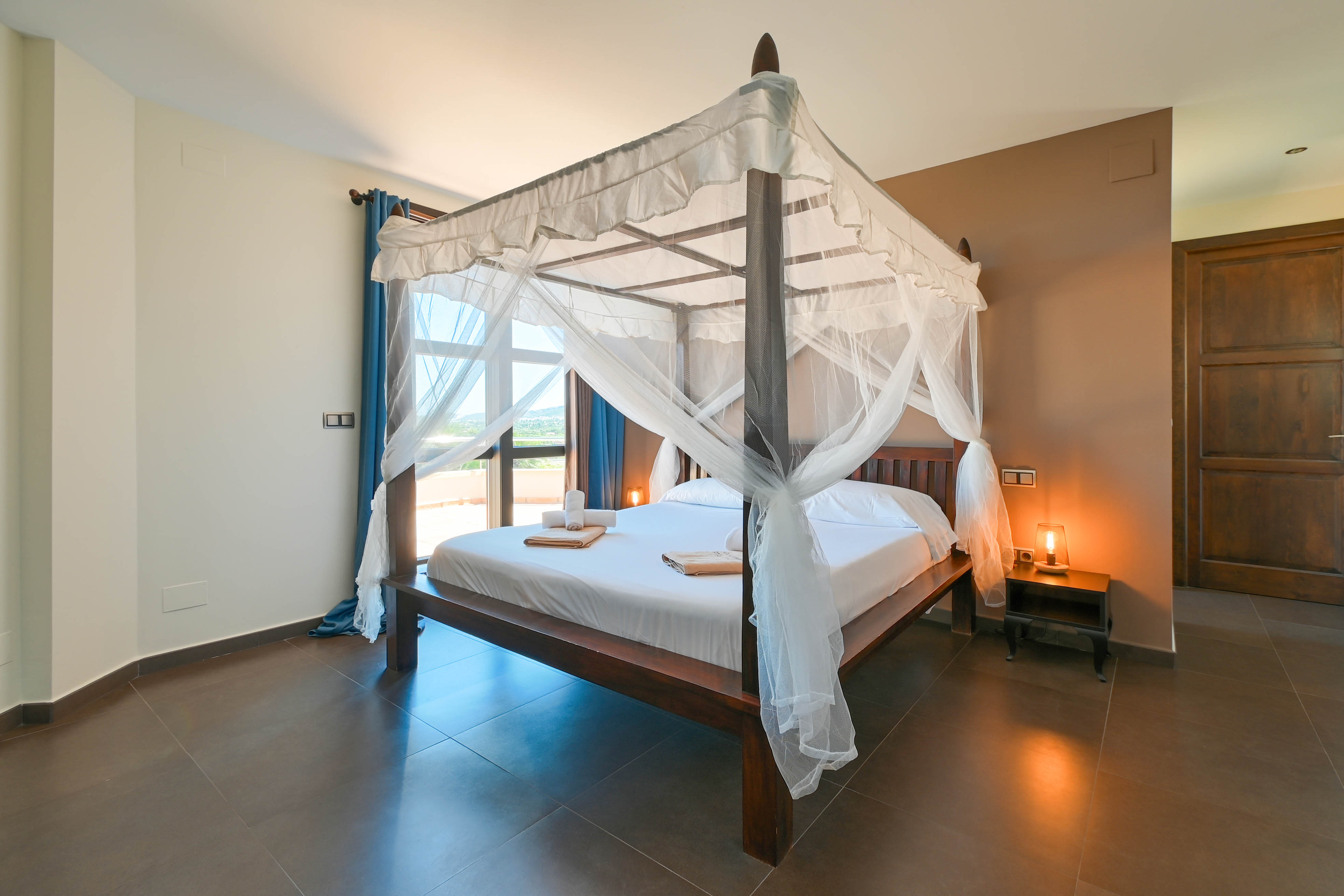 Dormitorio con cama con dosel de madera oscura y cortinas blancas, suelo de baldosas marrón.