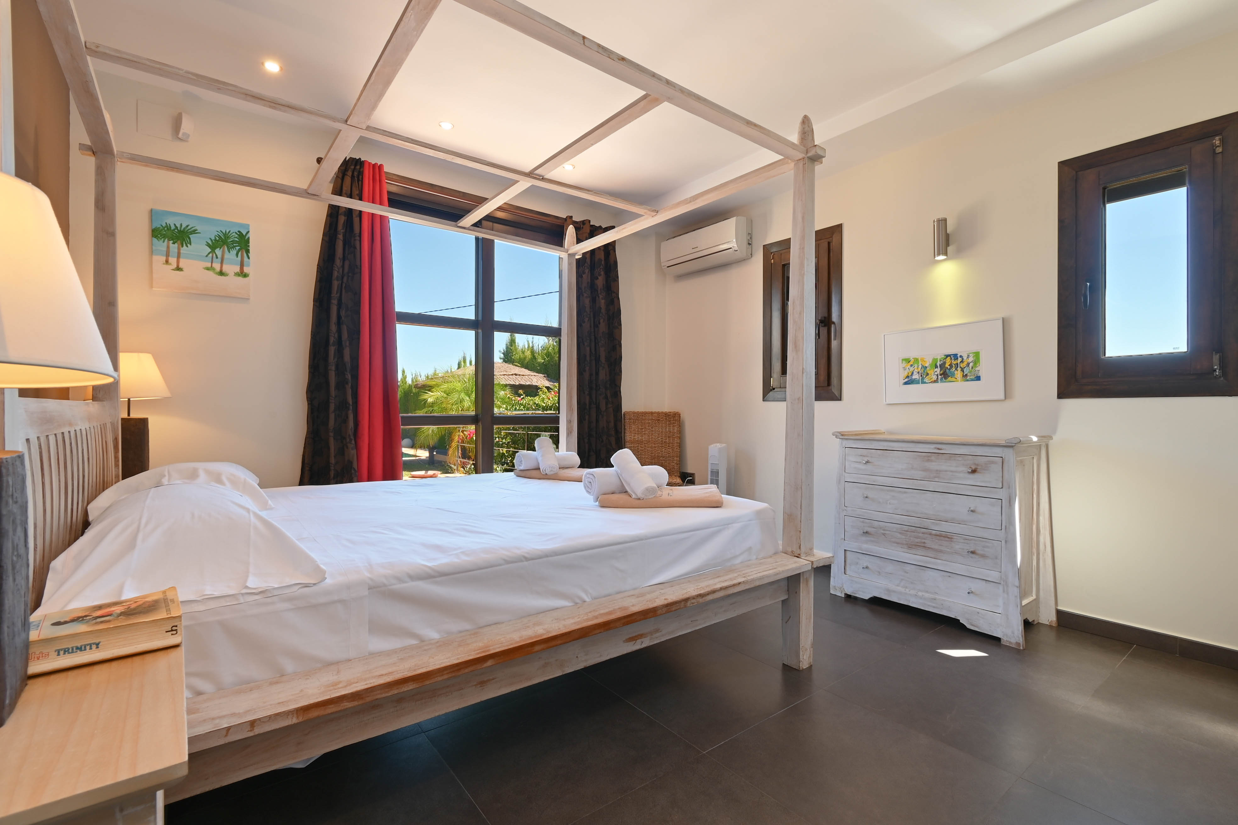 Dormitorio luminoso con cama con dosel de madera clara y cómoda blanca decapada.