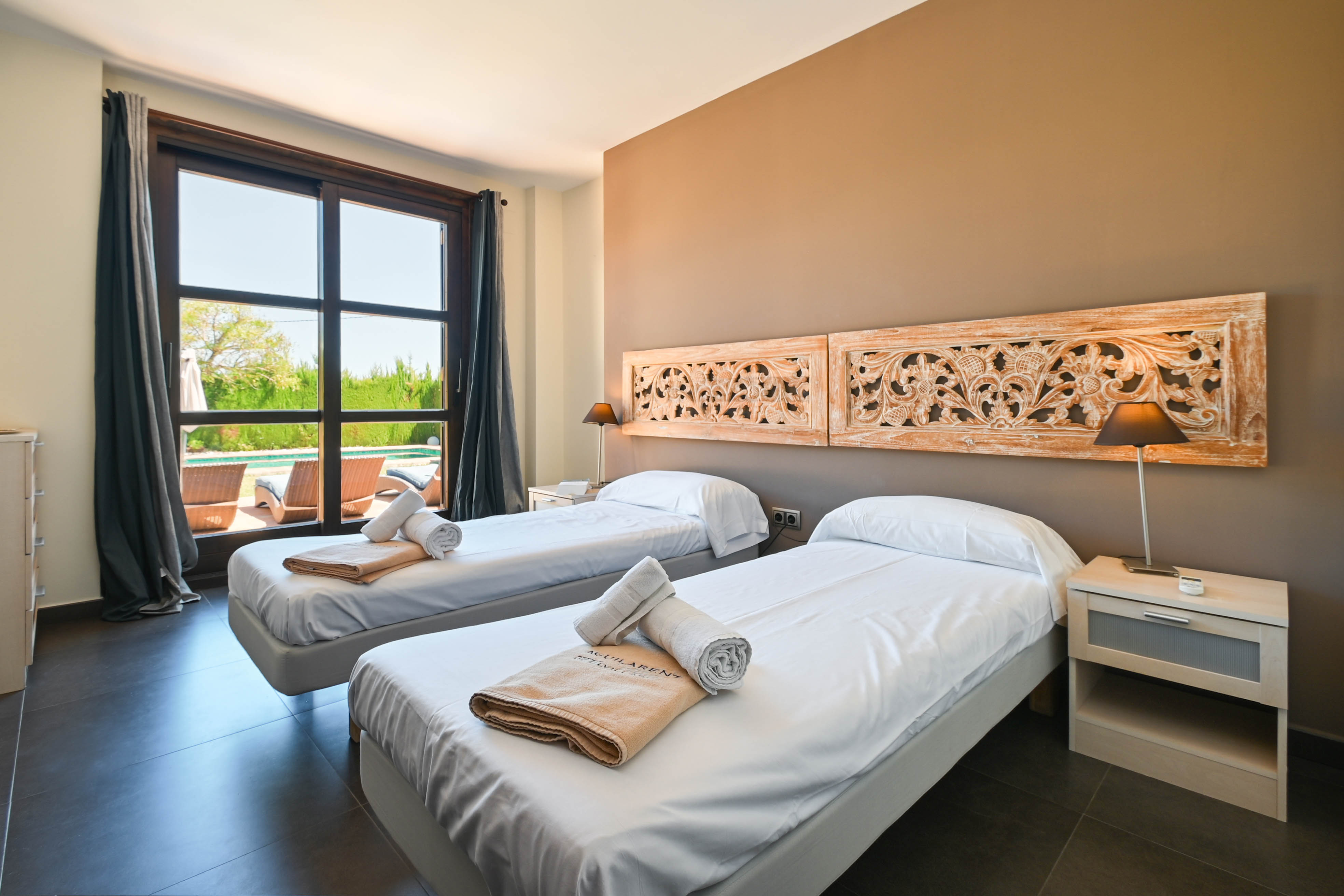 Dormitorio luminoso con dos camas individuales, cabeceros de madera tallada y vistas a la piscina.