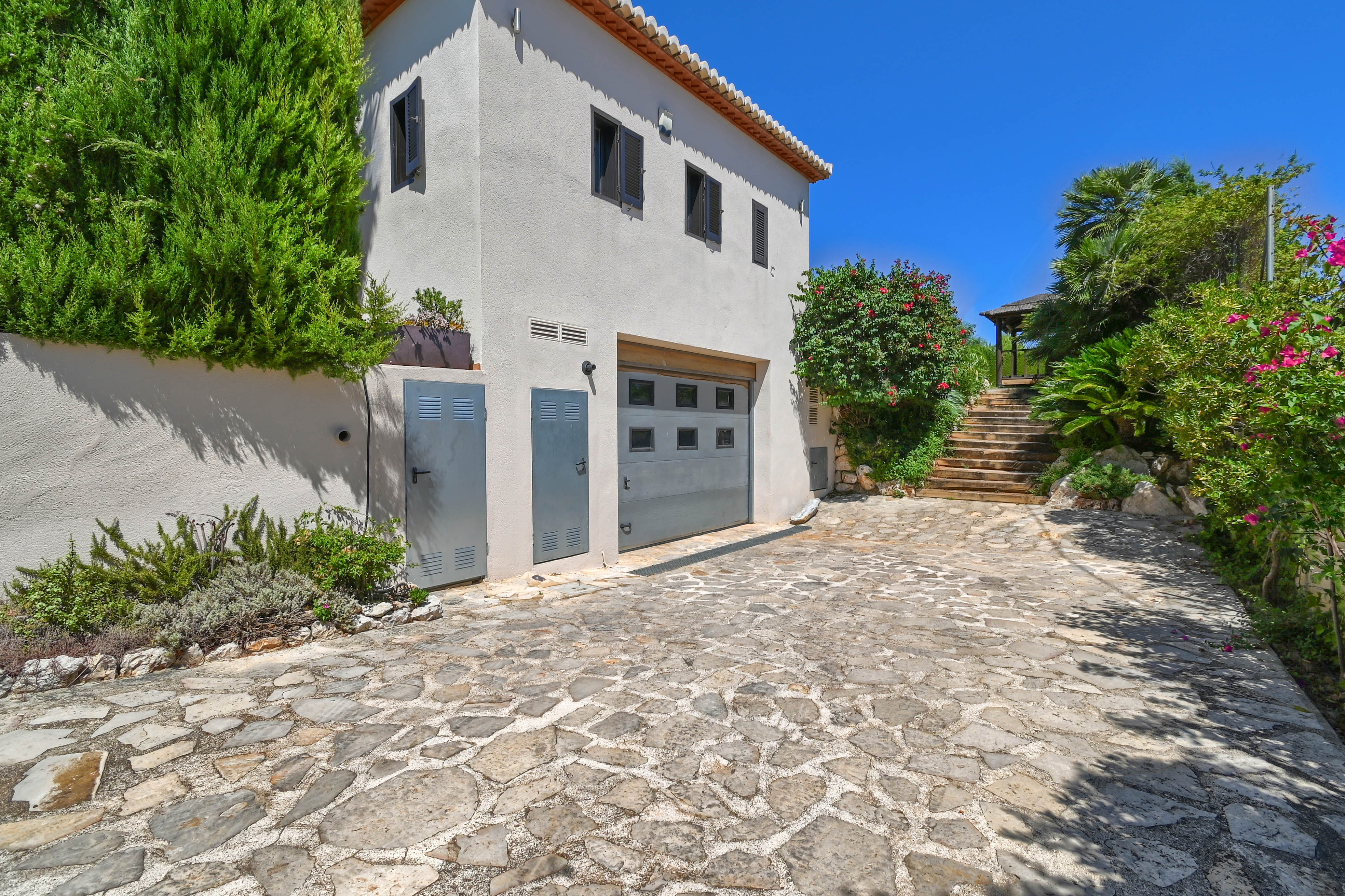 Exterior de la villa con camino de piedra, vegetación y garaje gris.