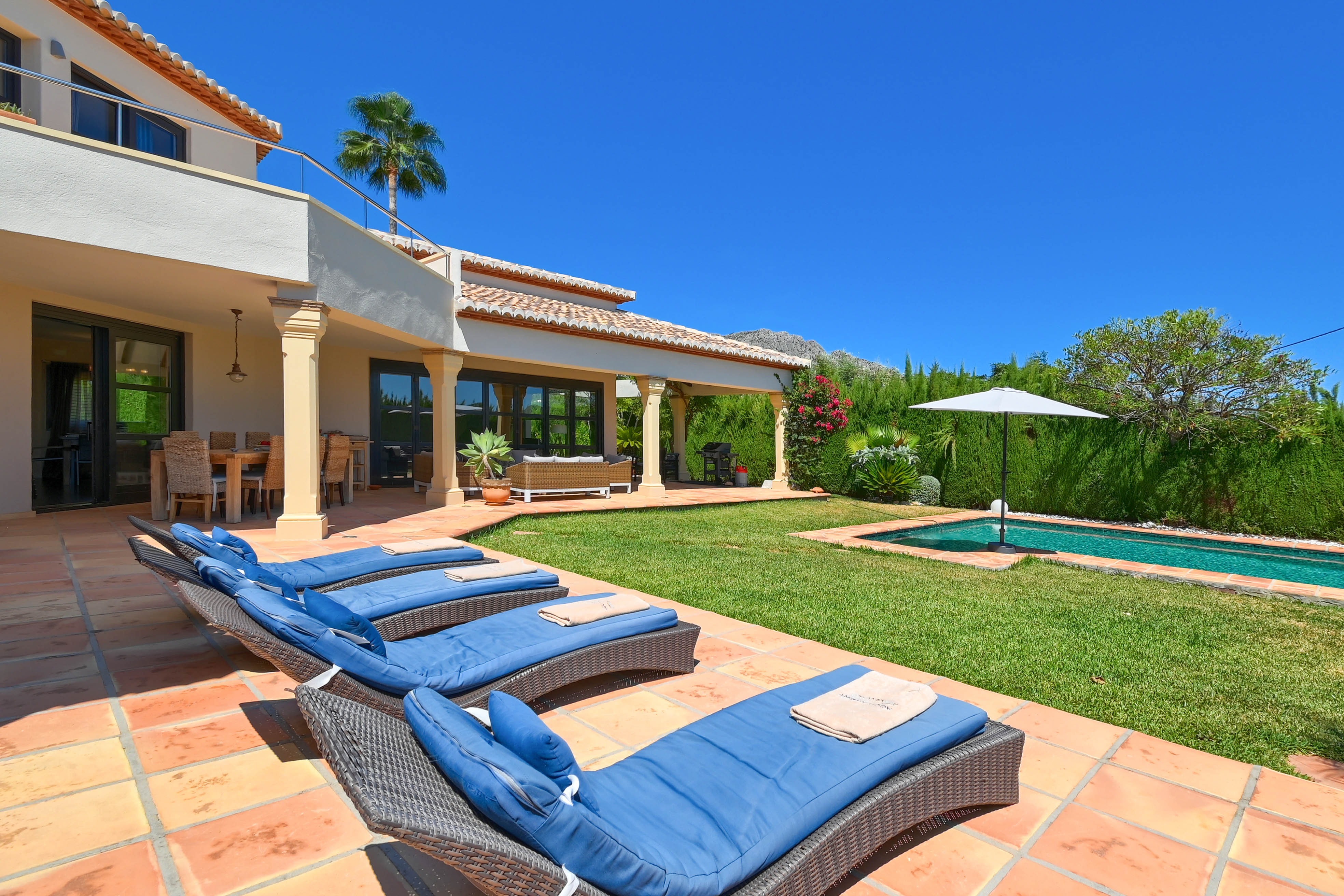 Terraza con tumbonas azules y piscina alargada en jardín con césped.