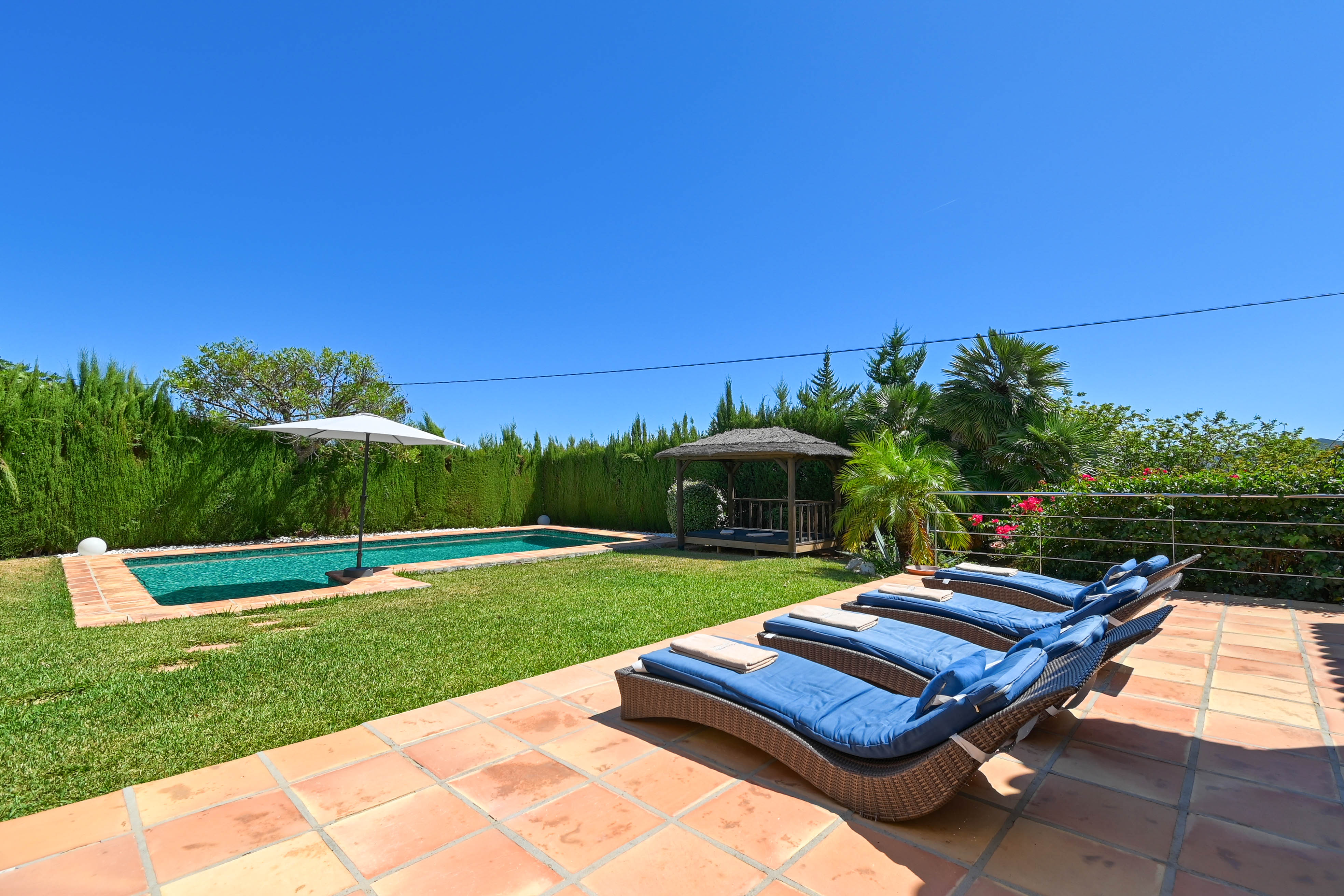 Terraza solárium con tumbonas de ratán azul, piscina y cenador en jardín con césped.