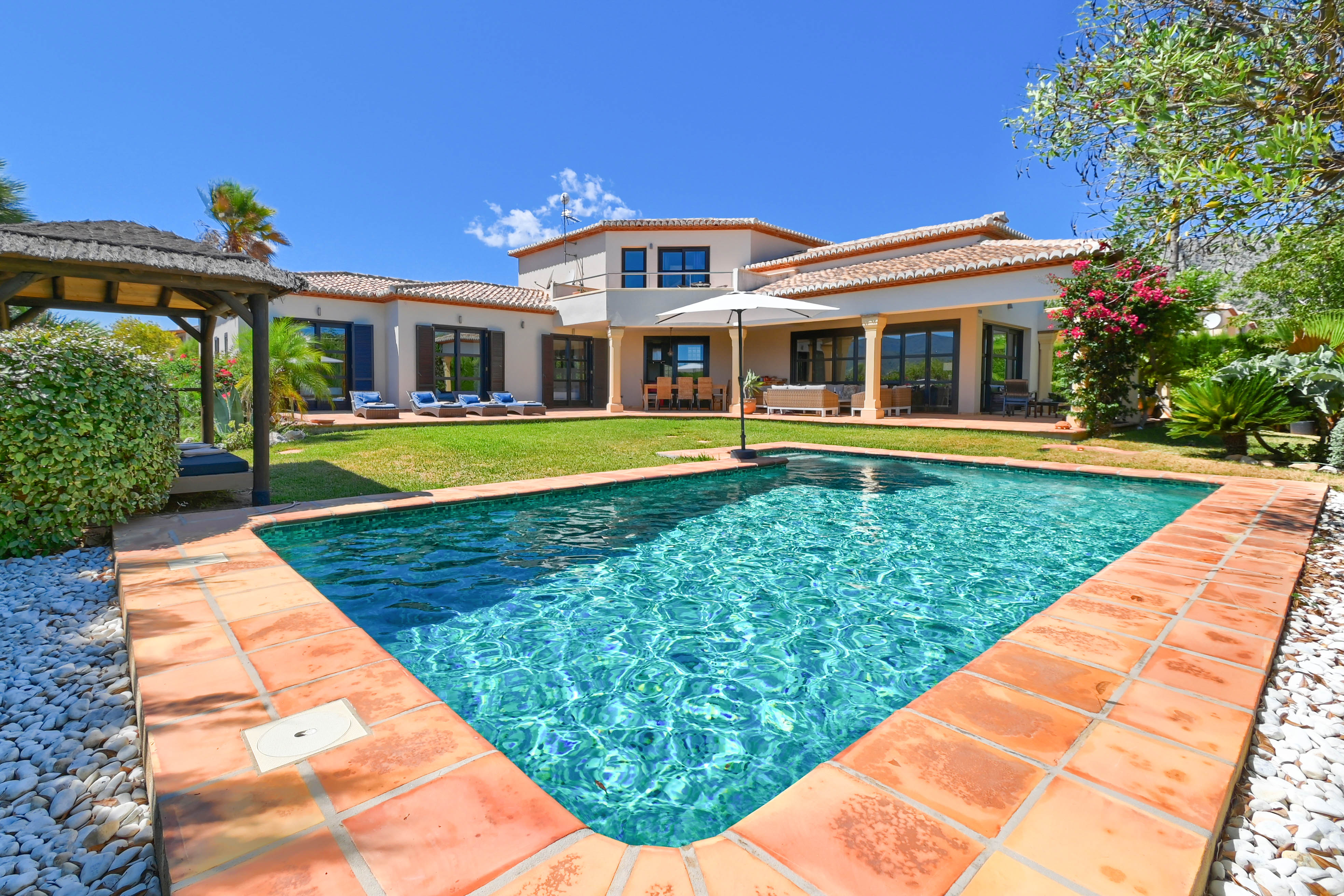 Vista de la piscina turquesa con borde de terracota y la villa con jardín y cenador.