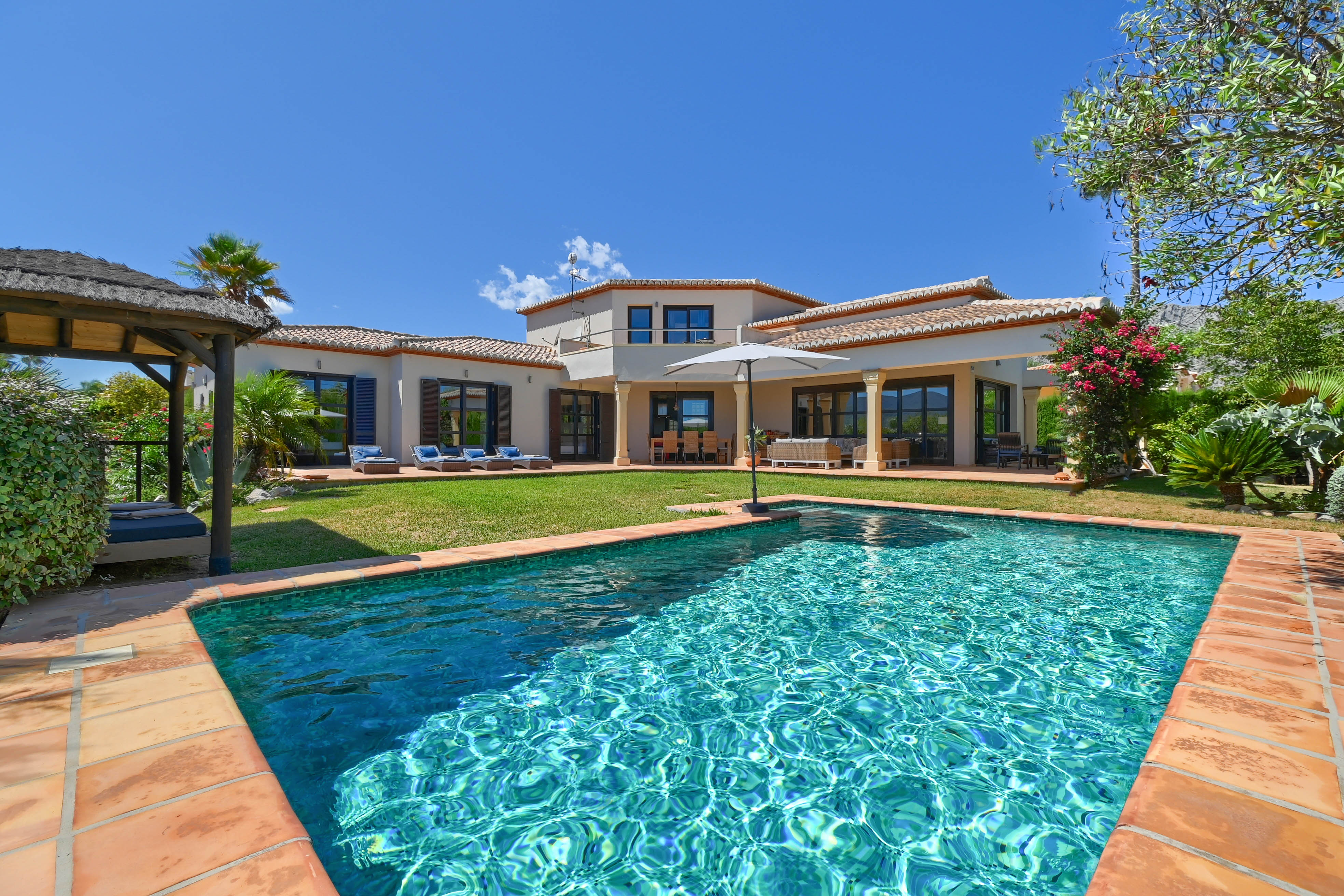Vista de la piscina turquesa con borde de terracota y la villa de dos pisos con contraventanas.