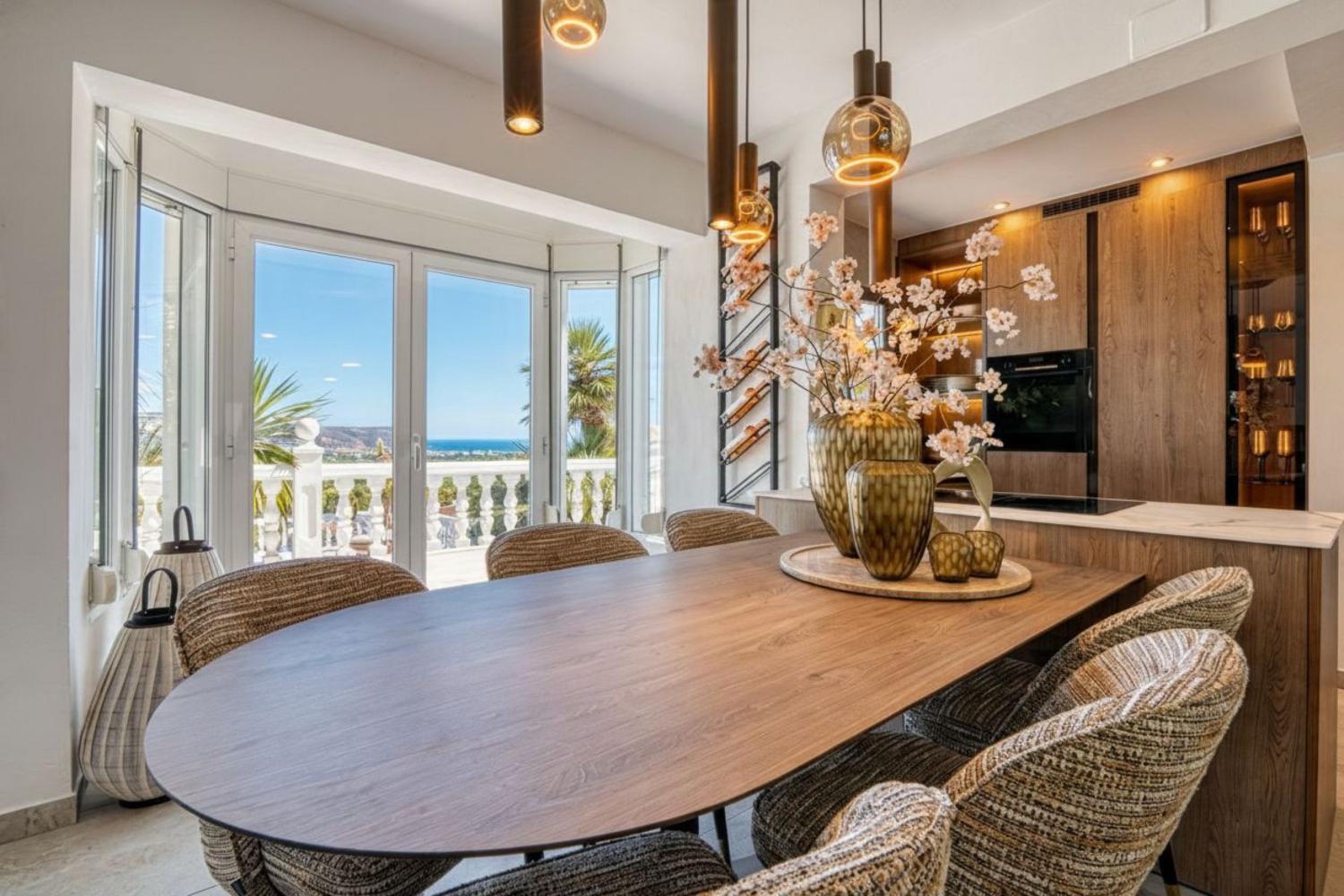 Comedor moderno con mesa de madera, sillas tapizadas y vistas al mar Mediterráneo a través de amplios ventanales.