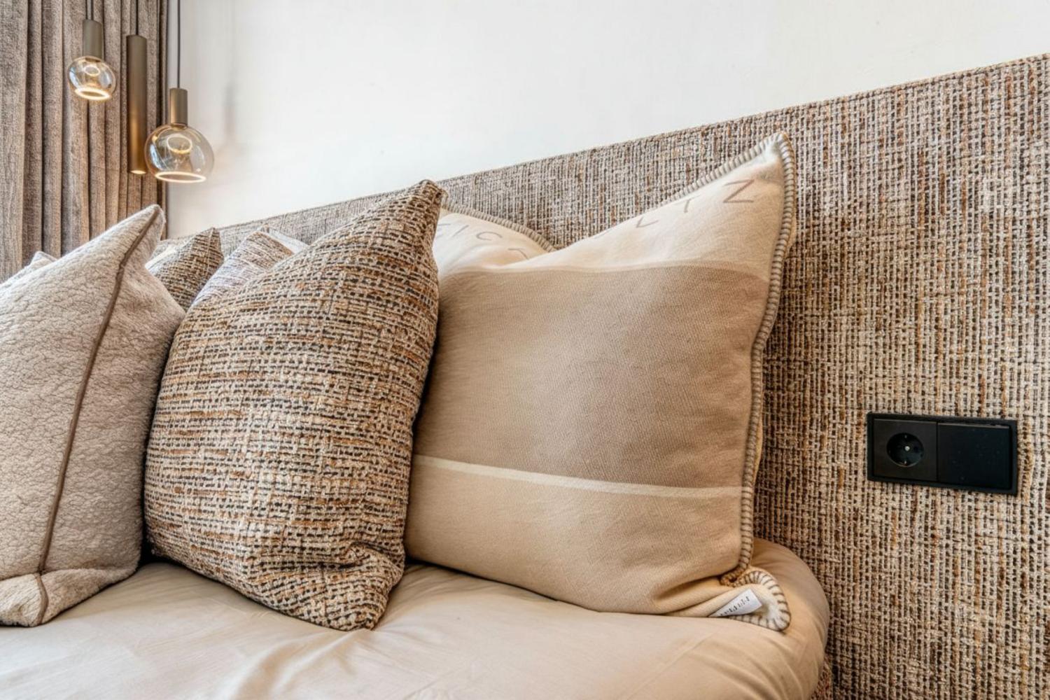 Detalle de la cama con cabecero de textura tejida y cojines decorativos en tonos neutros. Enchufe integrado en el cabecero.