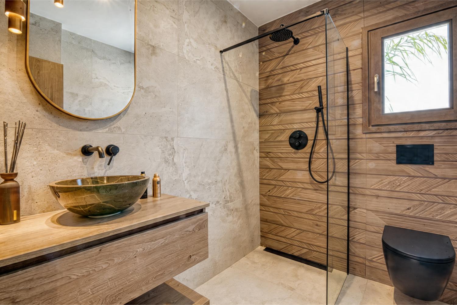 Detalle del baño con lavabo de piedra sobre encimera de madera y ducha con mampara de cristal.