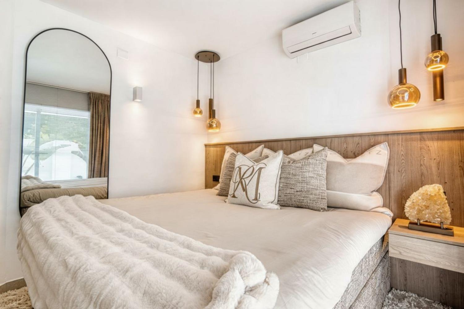 Dormitorio con cabecero de madera, iluminación colgante dorada y espejo de cuerpo entero.