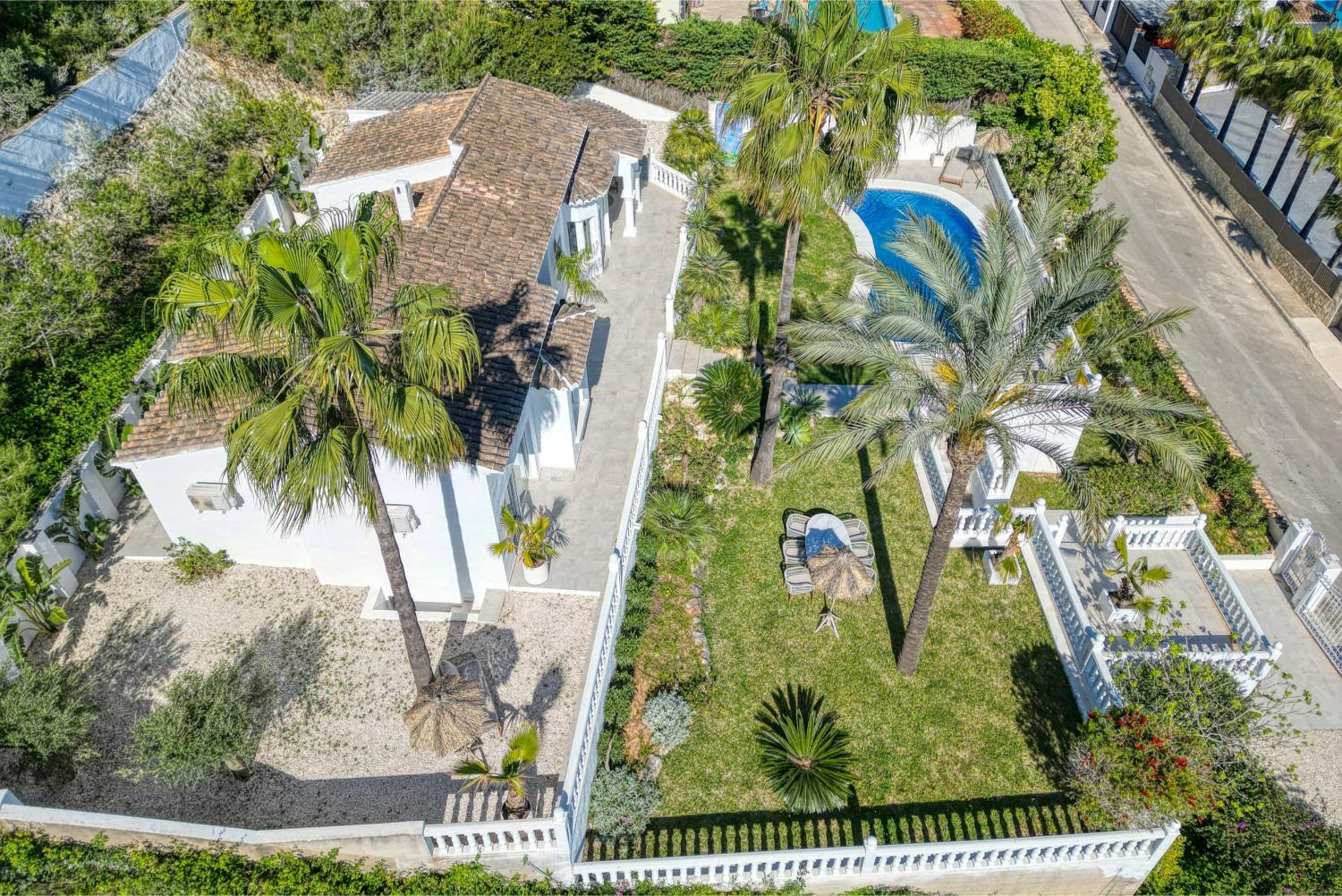 Vista aérea de la villa con jardín, piscina y terraza. Diseño mediterráneo con muros blancos y vegetación exuberante.