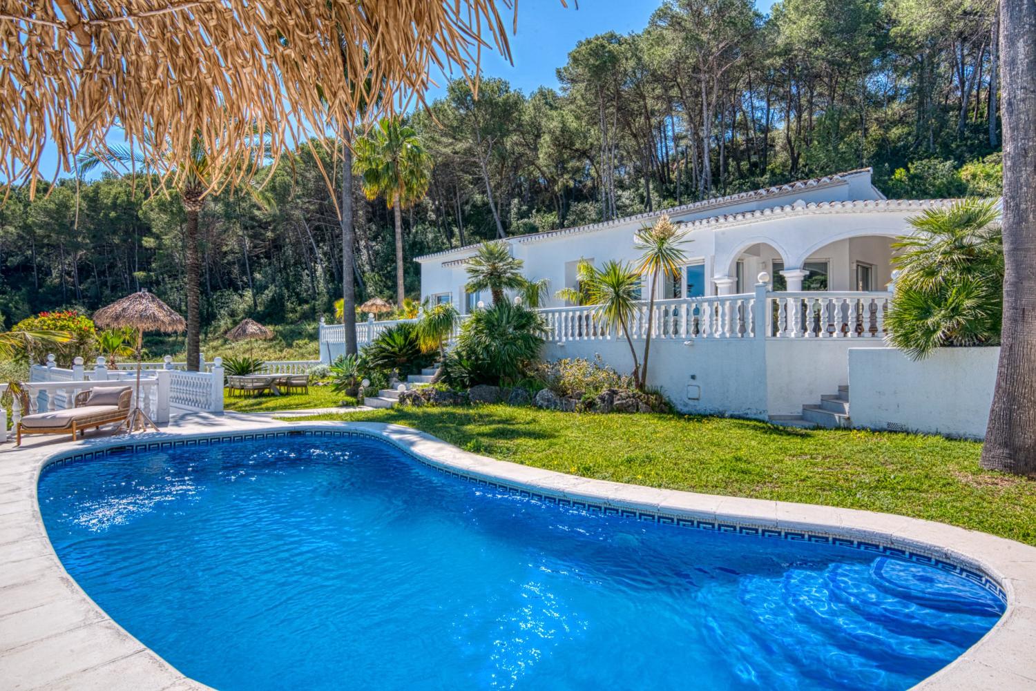 Vista de la piscina azul turquesa con borde de baldosas, rodeada de césped y vegetación exuberante, con la villa blanca al fondo.