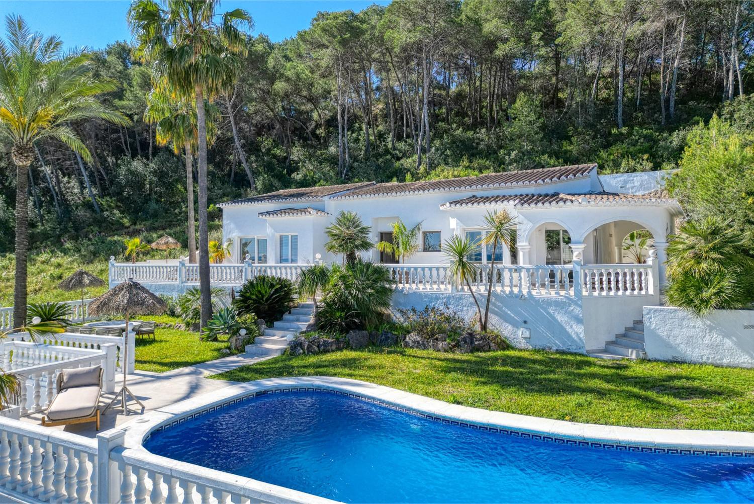 Vista de la villa desde la piscina, destacando su arquitectura mediterránea blanca y el jardín exuberante.