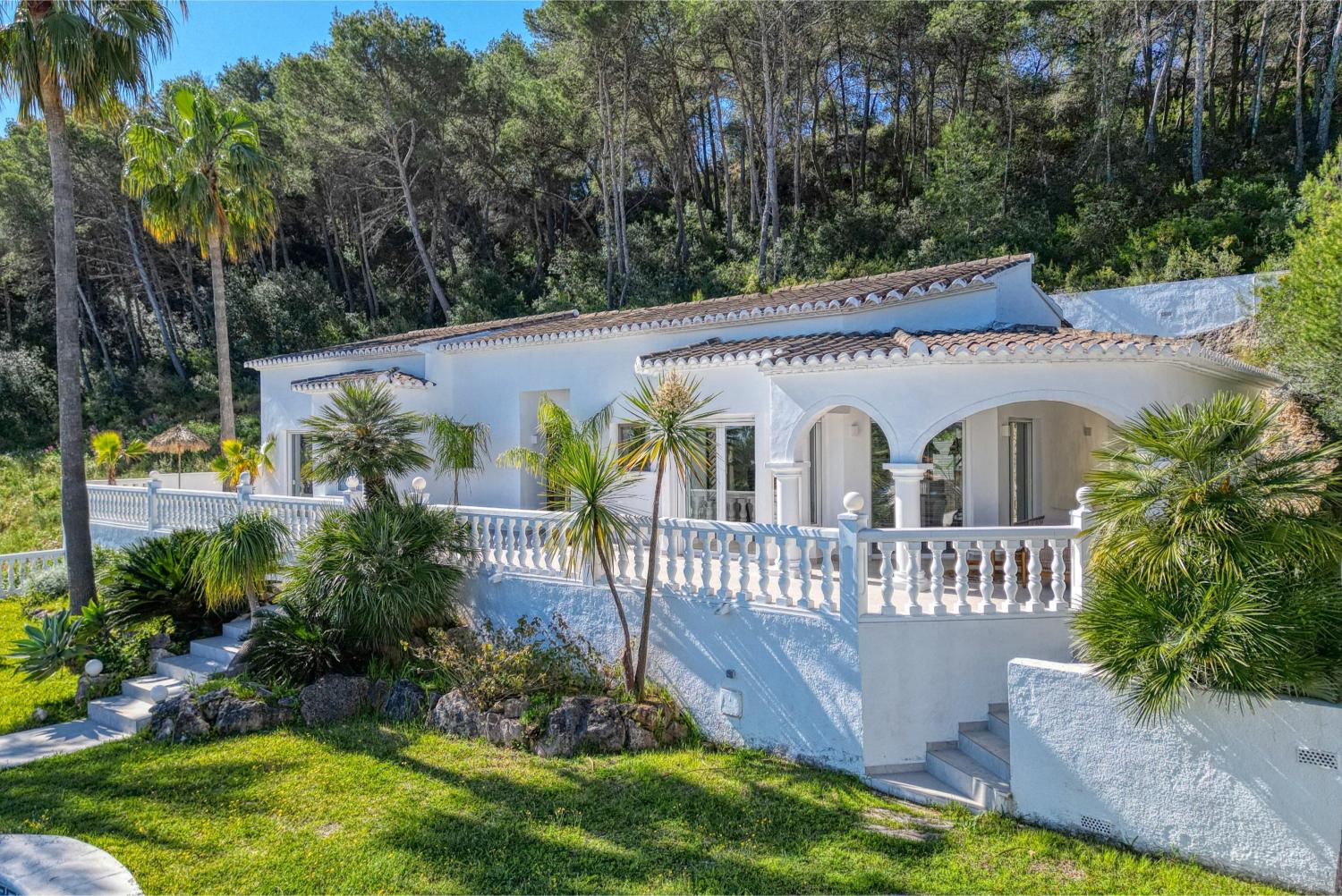 Vista exterior de la villa con terraza cubierta, barandilla de balaustres y jardín con vegetación mediterránea.