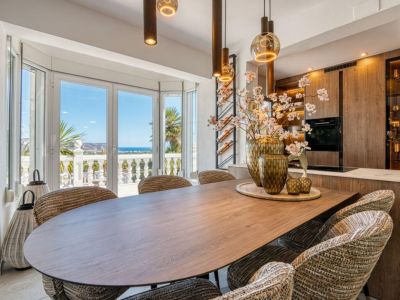 Comedor moderno con mesa de madera, sillas tapizadas y vistas al mar Mediterráneo a través de amplios ventanales.