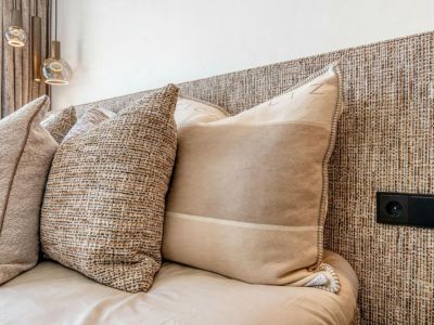 Detalle de la cama con cabecero de textura tejida y cojines decorativos en tonos neutros. Enchufe integrado en el cabecero.