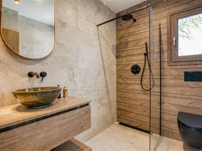 Detalle del baño con lavabo de piedra sobre encimera de madera y ducha con mampara de cristal.