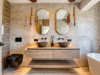 Elegante baño con lavabos de piedra, tocador flotante de madera y apliques dorados. Diseño moderno con azulejos neutros.