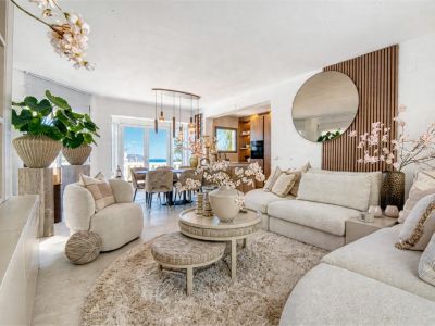 Salón luminoso con decoración neutra, alfombra de felpa y acceso al comedor con vistas al mar.