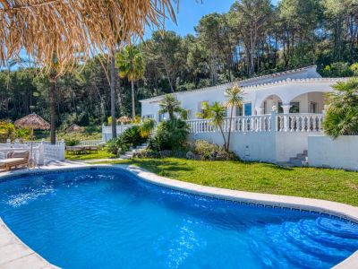 Vista de la piscina azul turquesa con borde de baldosas, rodeada de césped y vegetación exuberante, con la villa blanca al fondo.