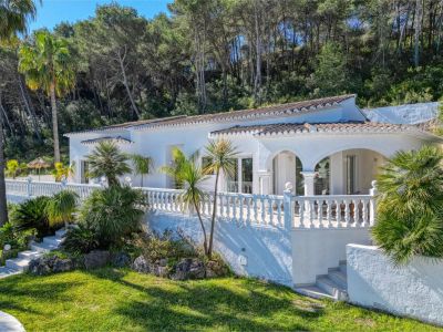 Vista exterior de la villa con terraza cubierta, barandilla de balaustres y jardín con vegetación mediterránea.