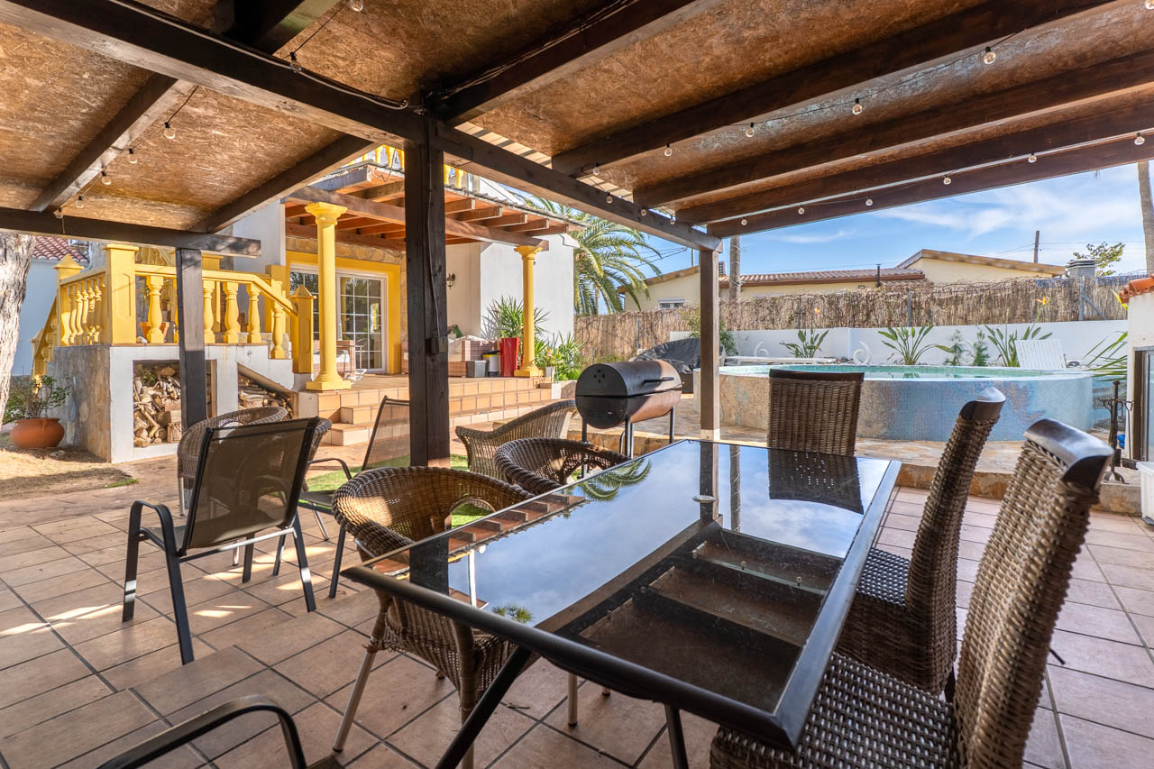 Comedor exterior cubierto con barbacoa y piscina en el jardín de la villa.