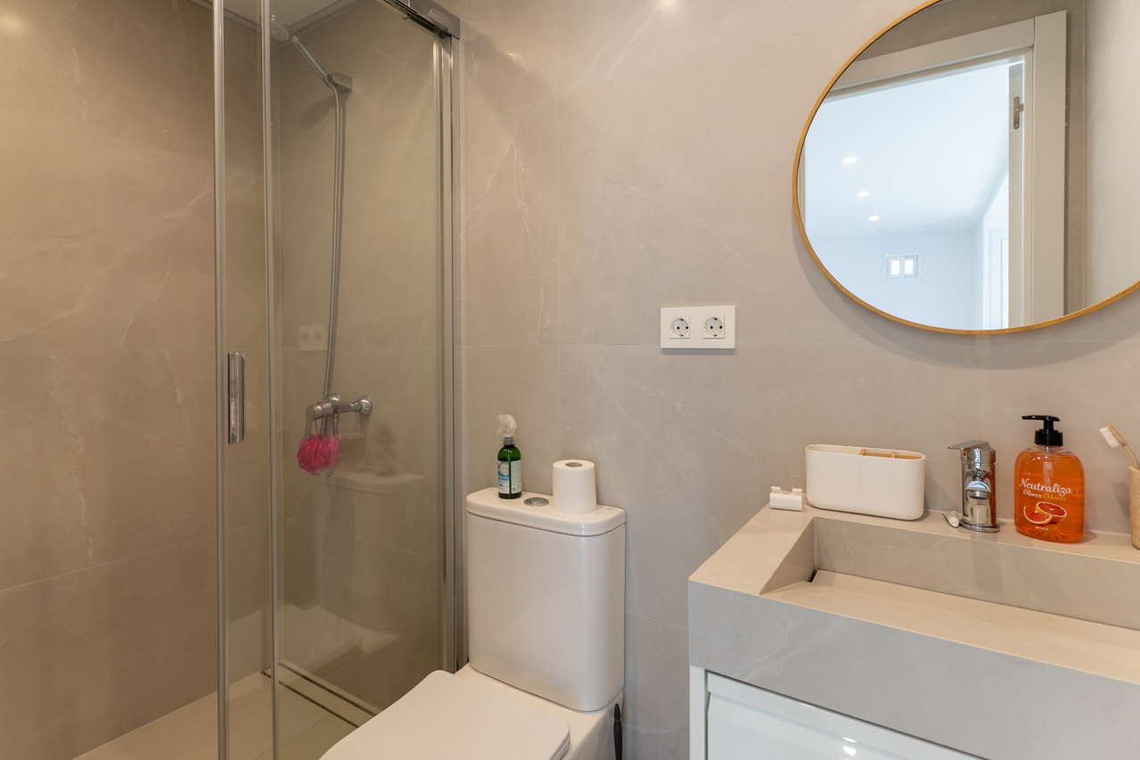 Detalle de baño con ducha de cristal, lavabo moderno y espejo circular con marco dorado.