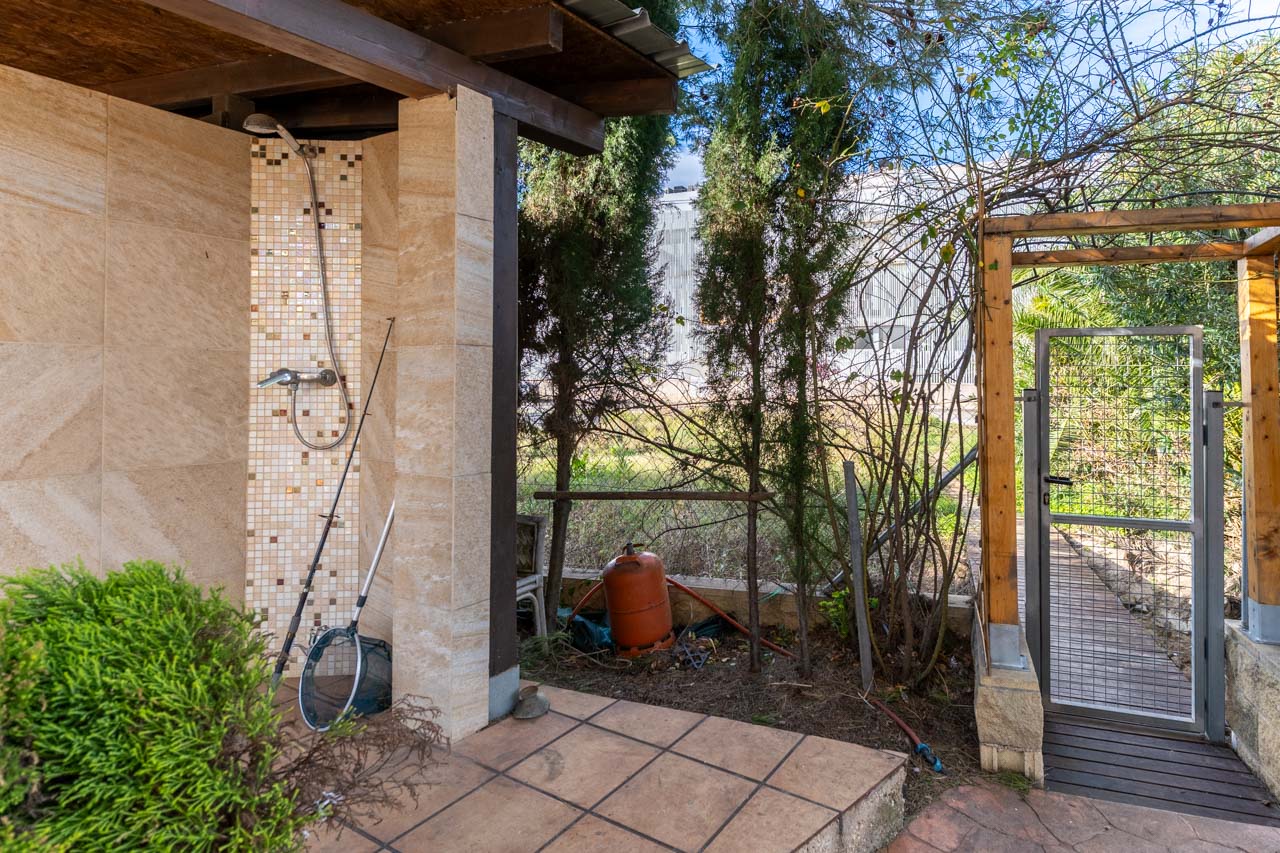 Detalle de ducha exterior con revestimiento cerámico y zona ajardinada con puerta metálica.