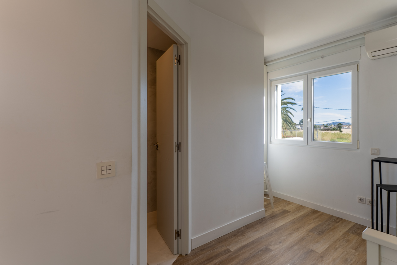 Habitación luminosa con suelo de madera y vistas al exterior. Puerta entreabierta al baño.