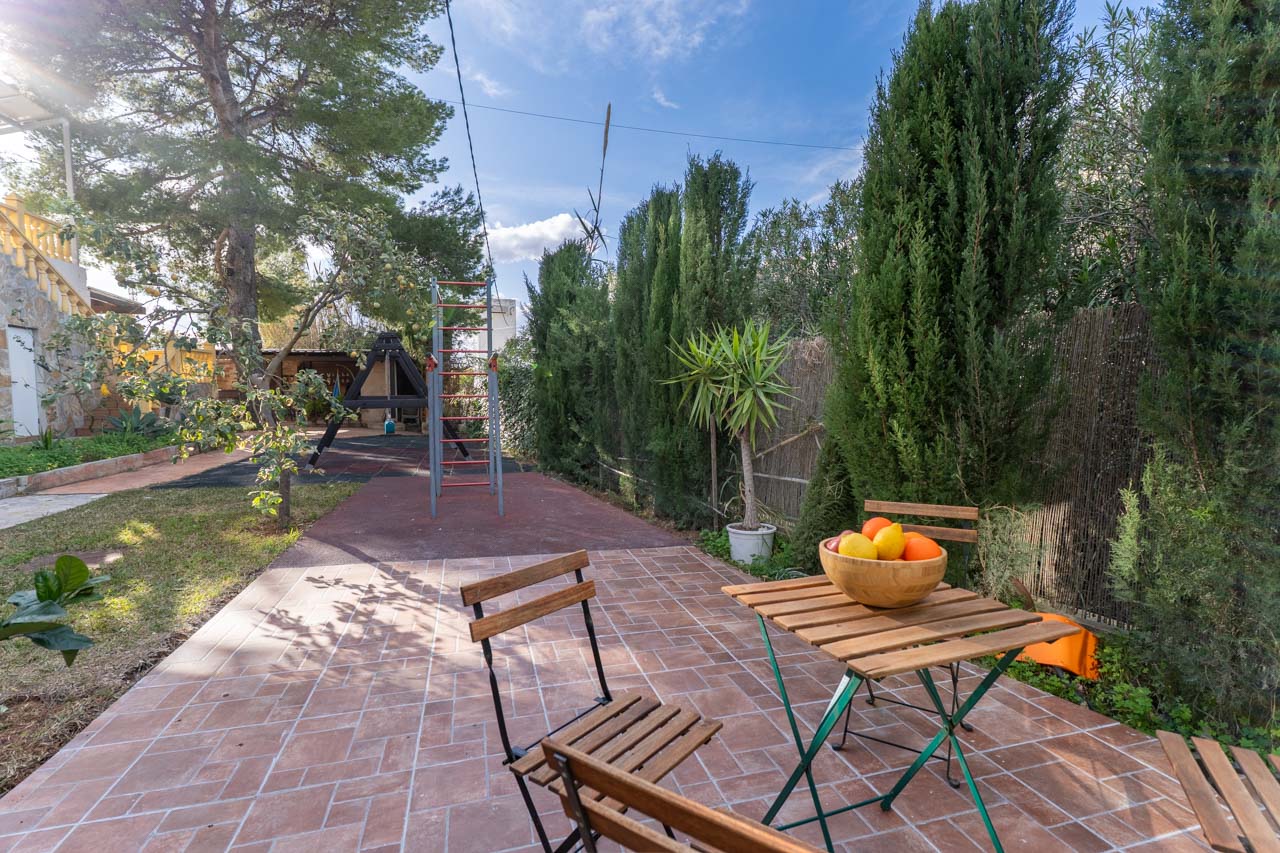 Terraza con mesa de madera y frutero, rodeada de vegetación y zona de juegos infantiles.