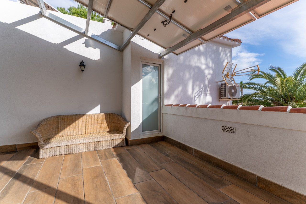 Terraza cubierta con suelo cerámico y sofá de mimbre, iluminada por luz natural y con acceso al exterior.