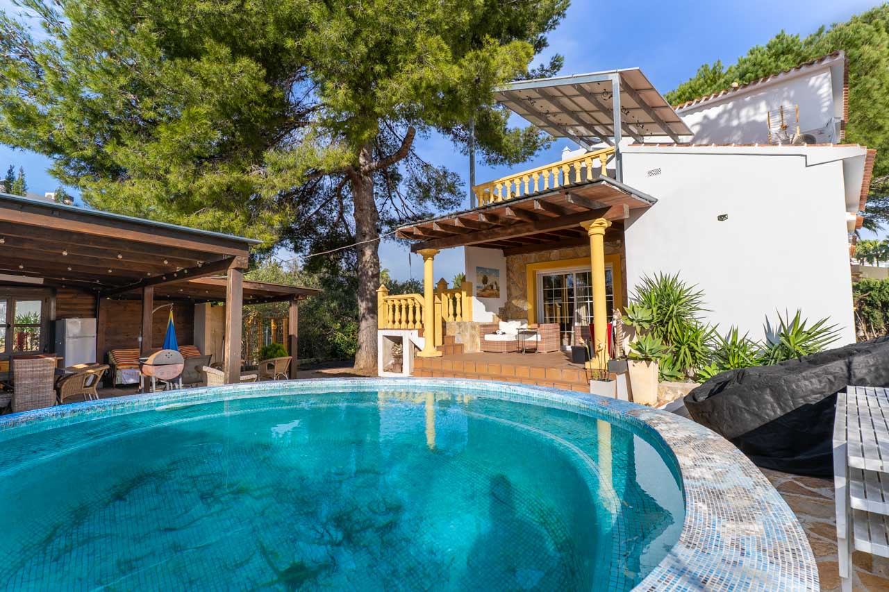 Vista de la piscina con mosaicos azules y la terraza cubierta con pilares amarillos.