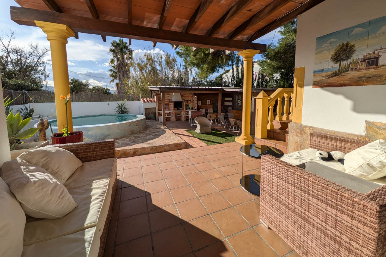 Vista del patio con piscina, barbacoa y zona de estar cubierta con muebles de ratán.