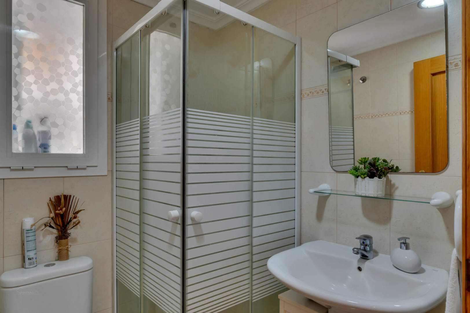 Detalle del baño con cabina de ducha esquinera y lavabo blanco con espejo rectangular.