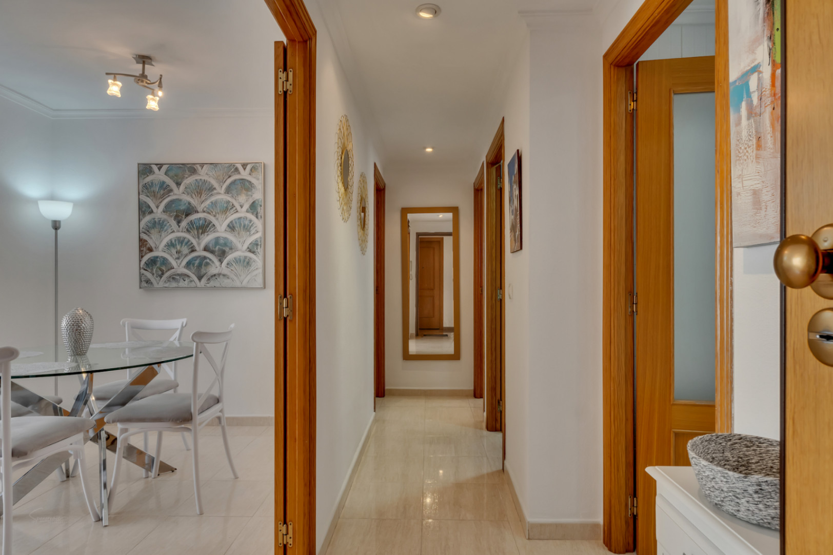 Detalle del pasillo con puertas de madera y suelo de baldosas color crema. Al fondo, espejo de cuerpo entero.