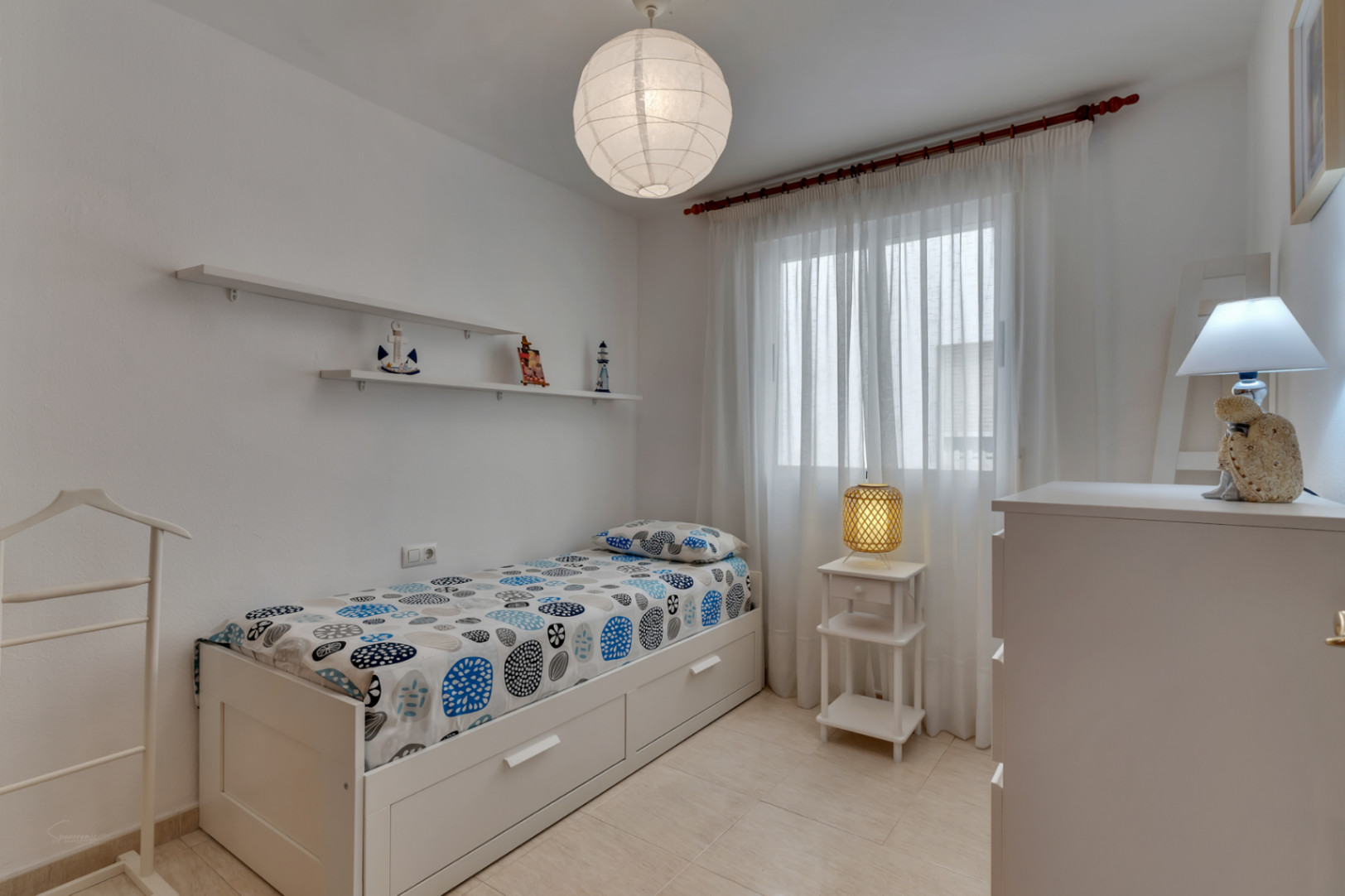 Habitación luminosa con cama individual, estantes decorativos y detalles náuticos.