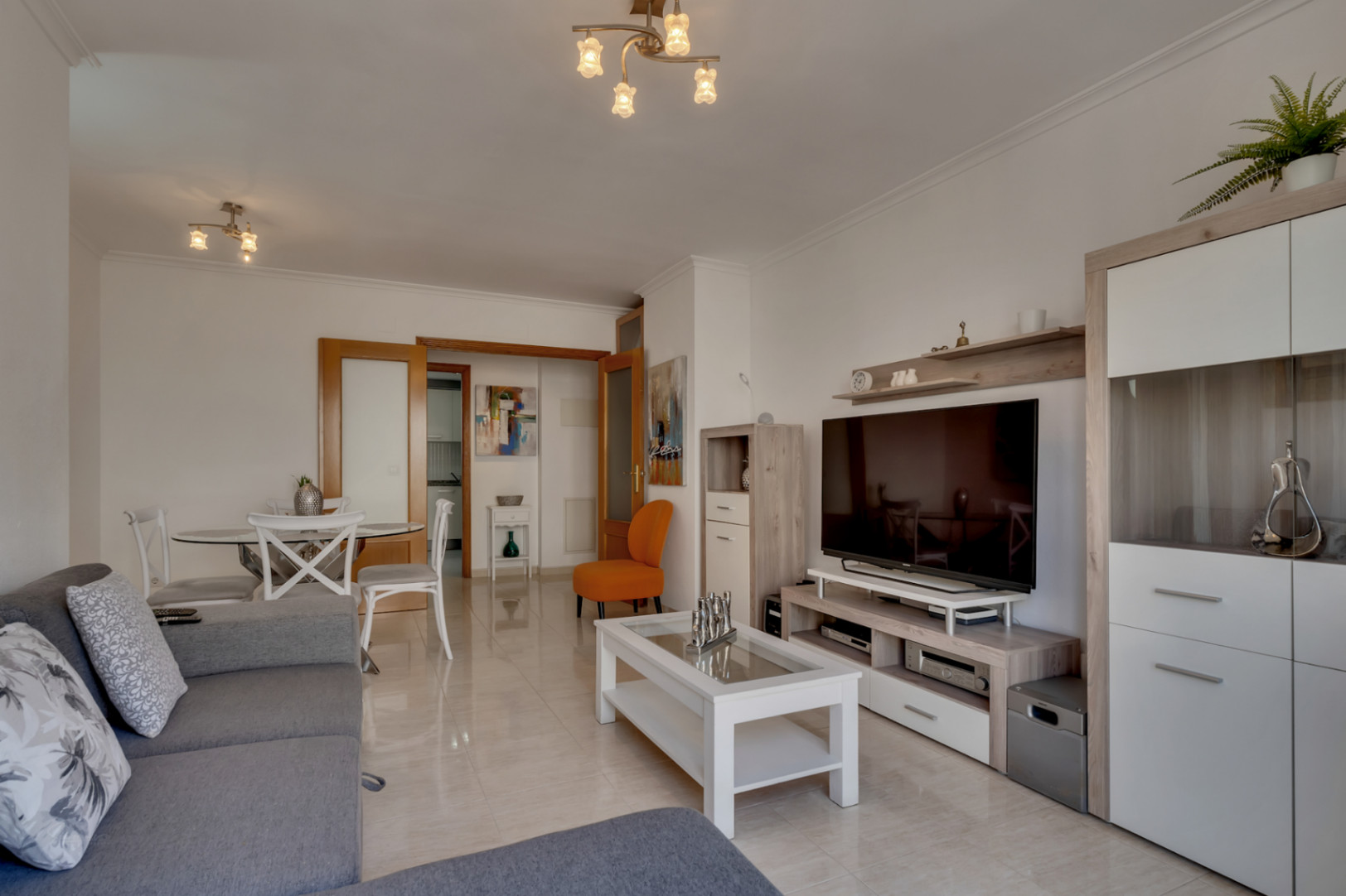 Salón luminoso con suelo de baldosas, sofá gris y mobiliario moderno en tonos neutros y blancos.