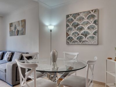 Comedor luminoso con mesa de cristal redonda y sillas blancas, complementado con arte moderno y una lámpara de pie.