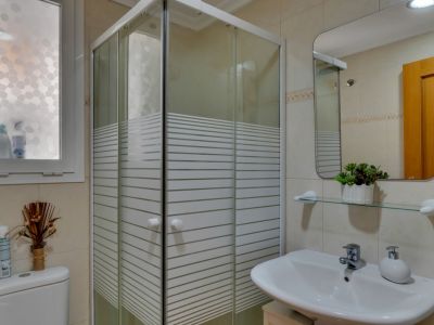 Detalle del baño con cabina de ducha esquinera y lavabo blanco con espejo rectangular.