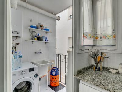 Detalle del lavadero con lavadora, pila y estanterías de azulejo blanco. Ventana con cortinas estampadas.
