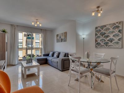 Salón-comedor luminoso con mesa de cristal, sofá gris esquinero y acceso al balcón con vistas urbanas.