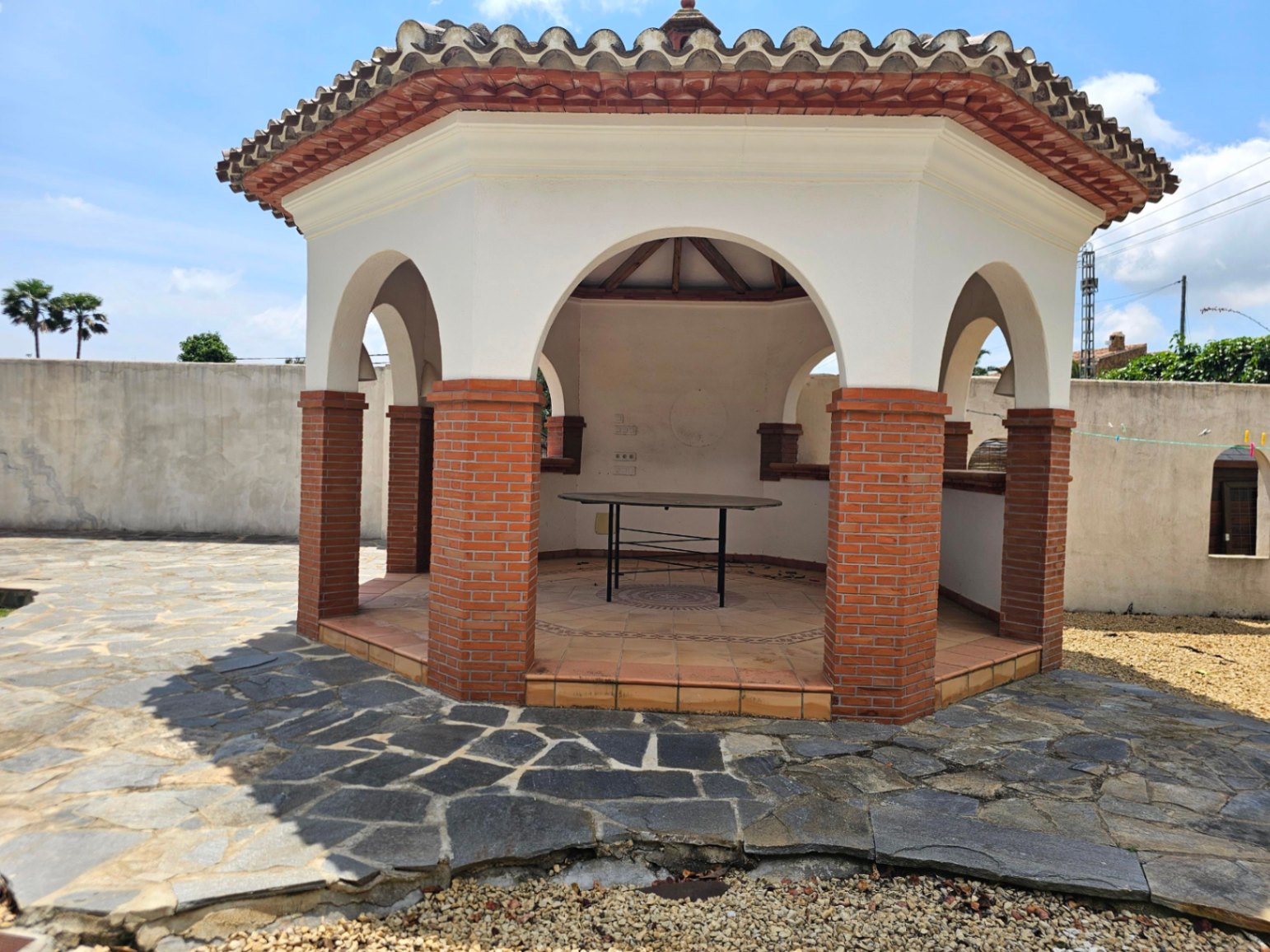 Cenador exterior con columnas de ladrillo y suelo de piedra irregular.